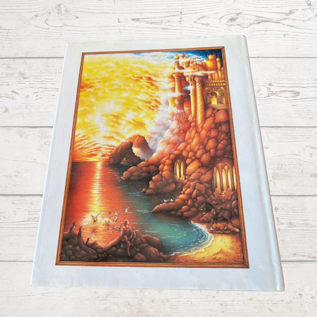 A book cover depicting a glowing golden castle beside a turquoise sea at sunset, photographed from above on a light wood plank surface
