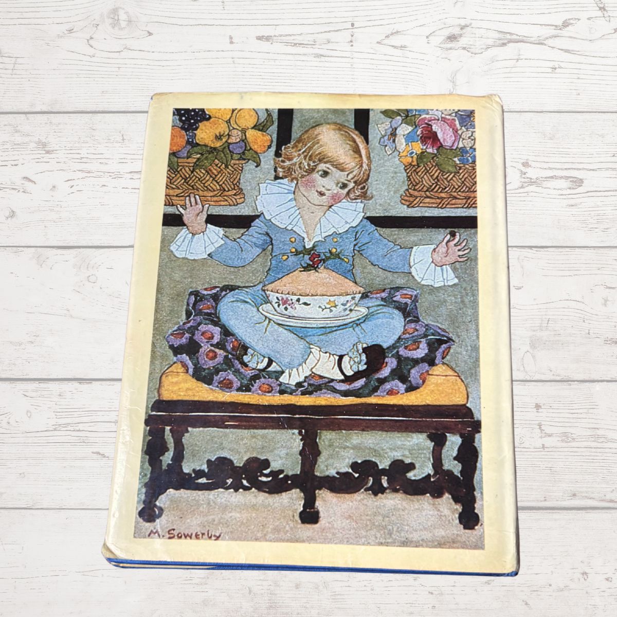 A vintage illustrated book o cover featuring a painted illustration of a young child in a blue outfit sitting on a bench with a teapot and cup, displayed on a light wood plank background.