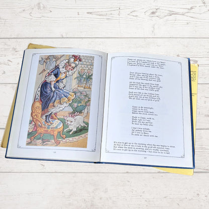 An open vintage children's book lying flat on a white wooden surface, showing a colorful illustration of a girl in a blue dress on the left page and printed text on the right page.