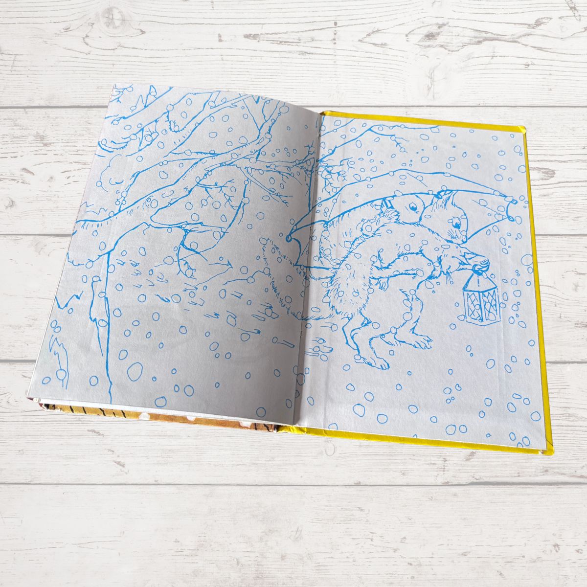 An open children's book with blue line-art illustrations of animals in a snowy scene, displayed on a light gray wood plank surface.