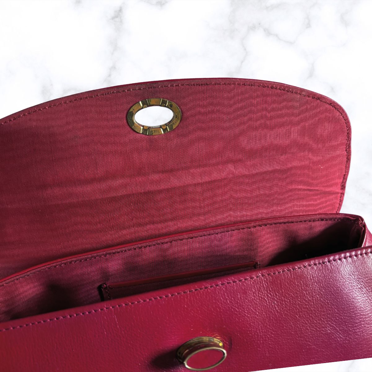 A close-up of a red  textured leather clutch bag showing the interior lining, a zippered compartment, and gold-tone metal hardware against a white marble background.