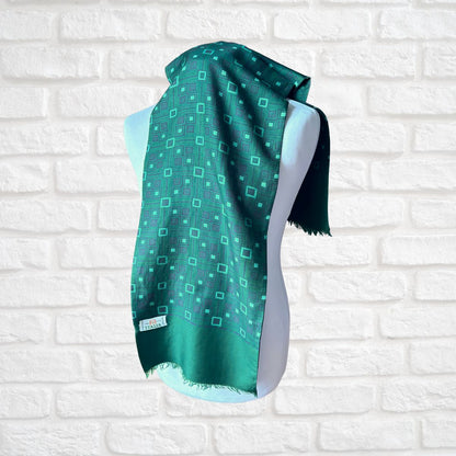 A teal green scarf with a geometric pattern of small squares and lines, draped on a white mannequin torso against a white brick wall background.