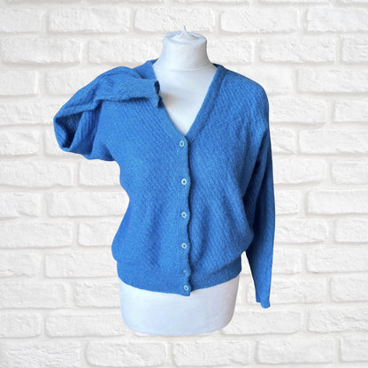 A bright blue textured knit cardigan sweater displayed on a white mannequin torso against a white painted brick wall background.