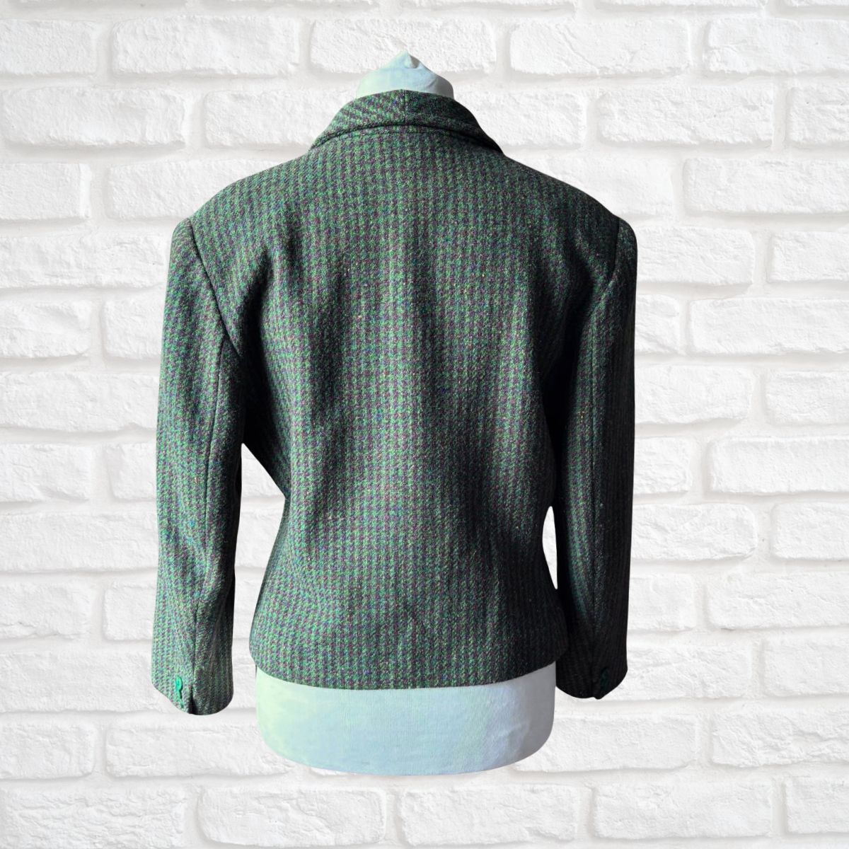 A green and brown textured tweed blazer jacket displayed on a white mannequin torso against a white brick wall background.