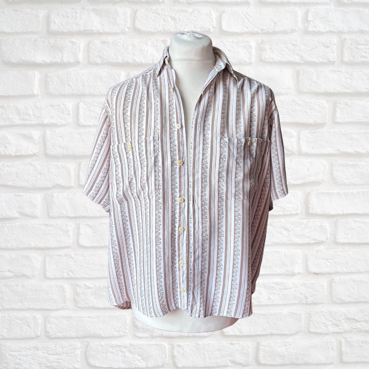 A short-sleeve button-up shirt with vertical stripes in beige, white, and light brown, displayed on a white mannequin torso against a white brick wall background.