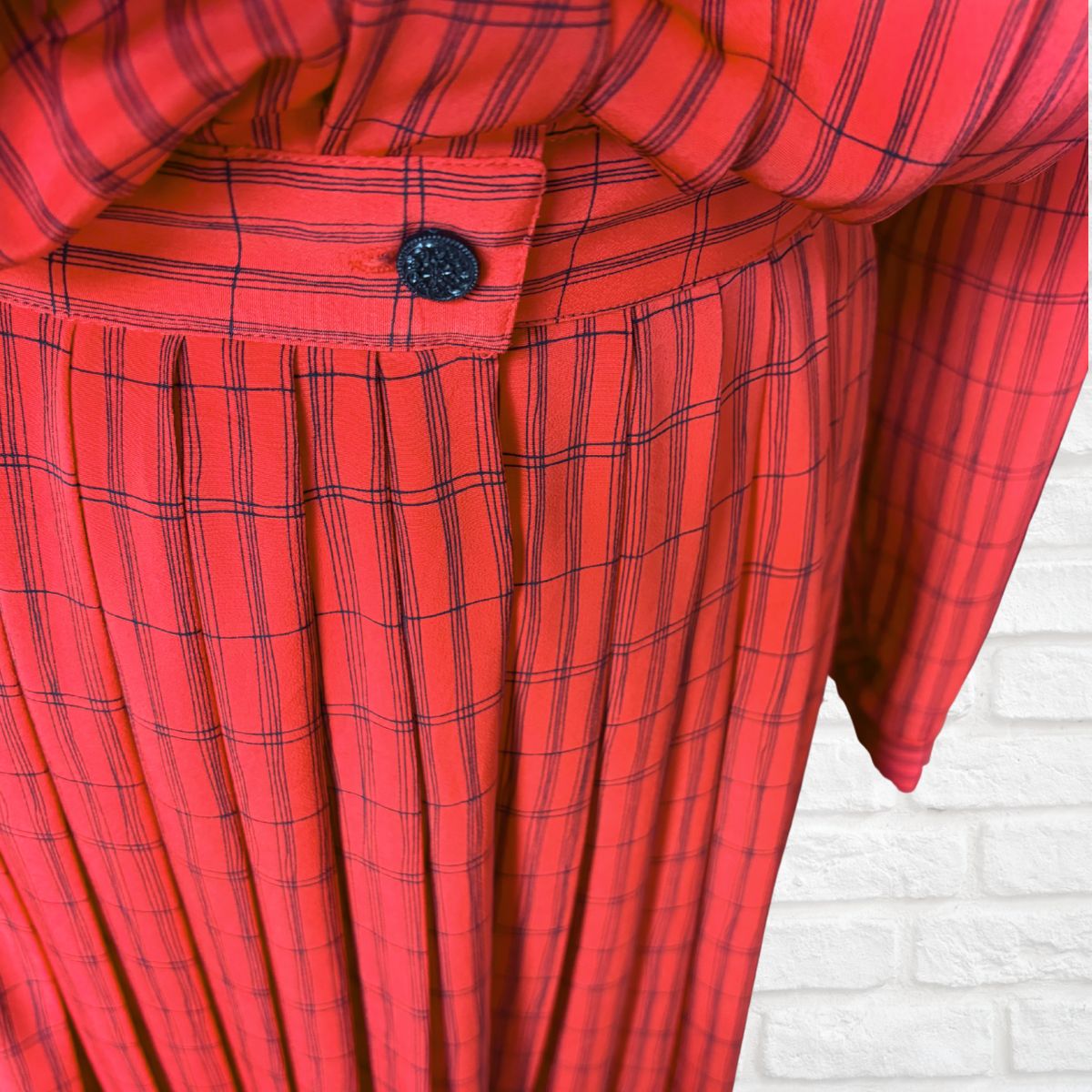 A close-up of a red and black plaid pleated dress, showing the waistband with a single black button and the pleated fabric texture, set against a white brick wall background.