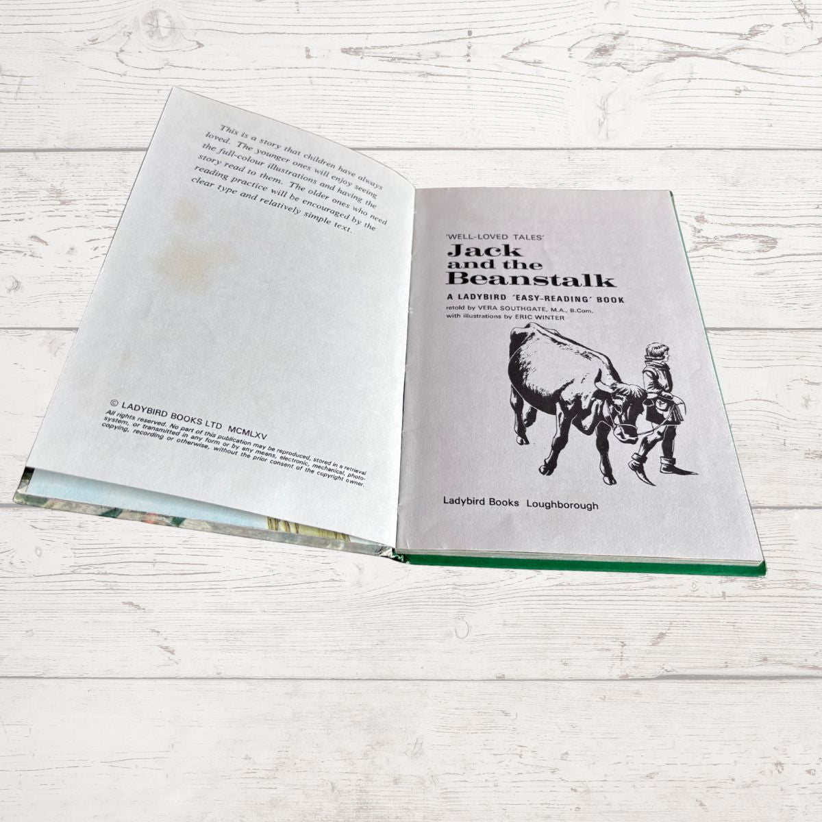 An open hardcover book titled 'Jack and the Beanstalk' laid flat on a white wood surface. The pages are light gray with black text and an illustration of a man climbing a beanstalk.