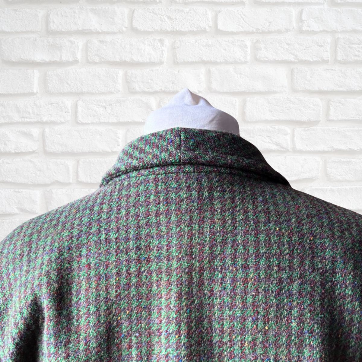 A close-up back view of a women's tweed jacket on a white mannequin torso, featuring a textured green and brown  houndstooth-like pattern against a white brick wall background