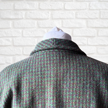 A close-up back view of a women's tweed jacket on a white mannequin torso, featuring a textured green and brown  houndstooth-like pattern against a white brick wall background