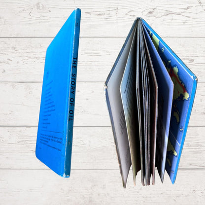 A vintage hardcover book titled 'THE STORY OF OIL' shown standing upright on its spine next to its open, fanned pages, set against a light whitewashed wood plank background.