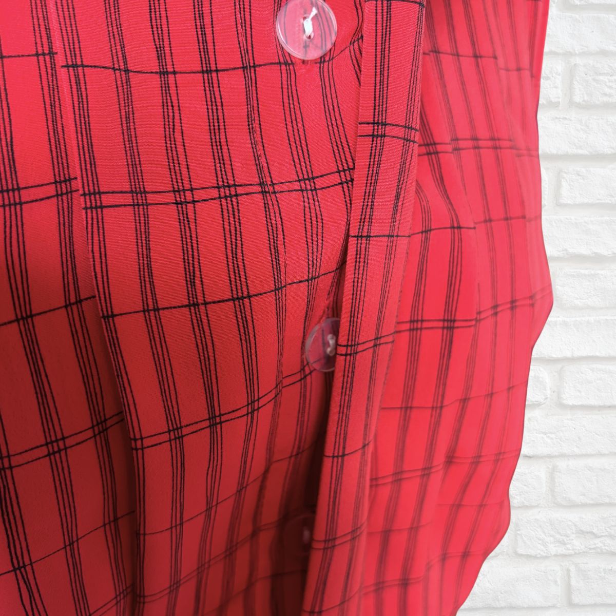 A close-up of a red dress ,featuring a black windowpane plaid pattern and translucent buttons, set against a white brick wall 