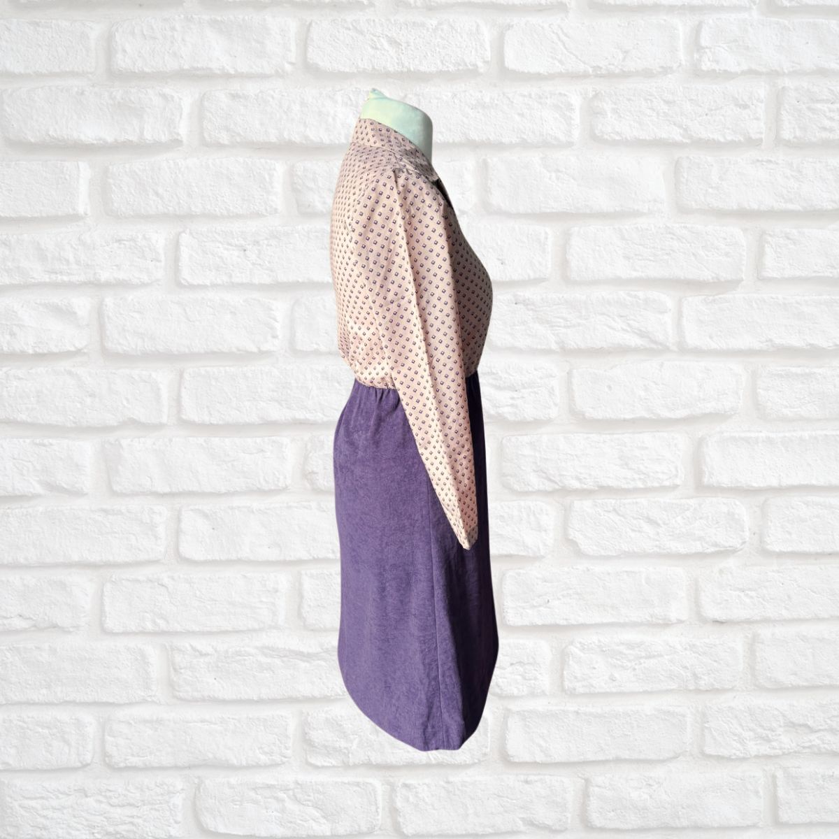 Mannequin wearing a dress with a beige top and purple skirt against a white brick wall.