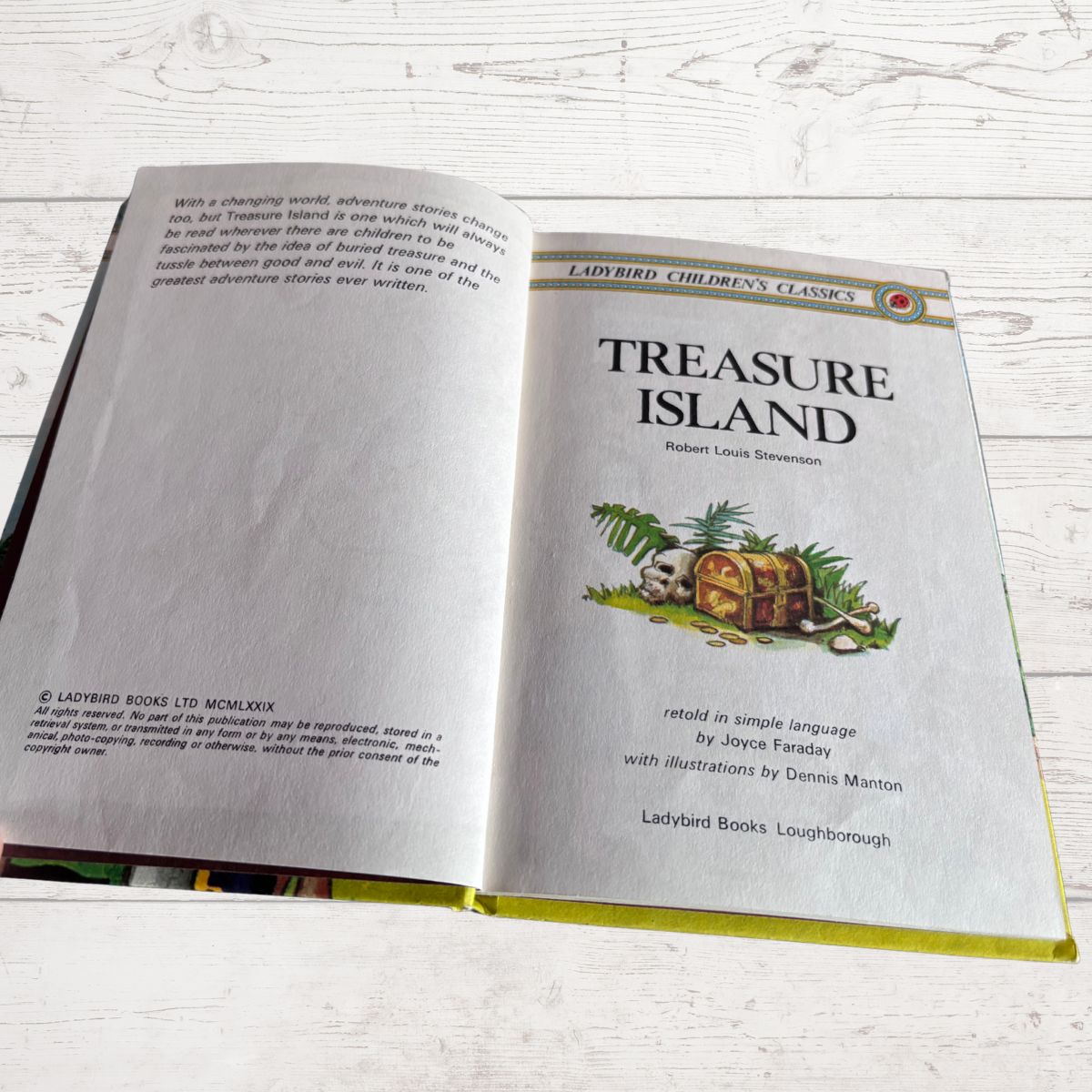 An open children's book titled 'Treasure Island' laid on a light whitewashed wooden surface. The left page shows story text, and the right page shows the title, author Robert Louis Stevenson, an illustration of a treasure chest with palm leaves, and Ladybird branding.