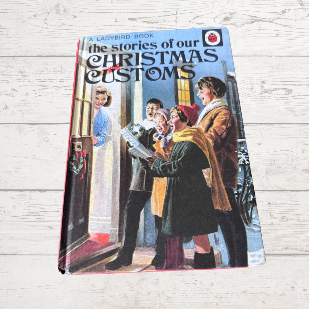 Vintage book titled 'The stories of our Christmas Customs' by Ladybird, shown on a light wood background. The cover features a colorful illustration of people singing in a doorway.