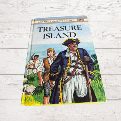 A vintage hardcover children's book titled 'Treasure Island' from Ladybird Children's Classics, featuring an illustration of pirates on the cover, photographed on a white wood-plank surface.