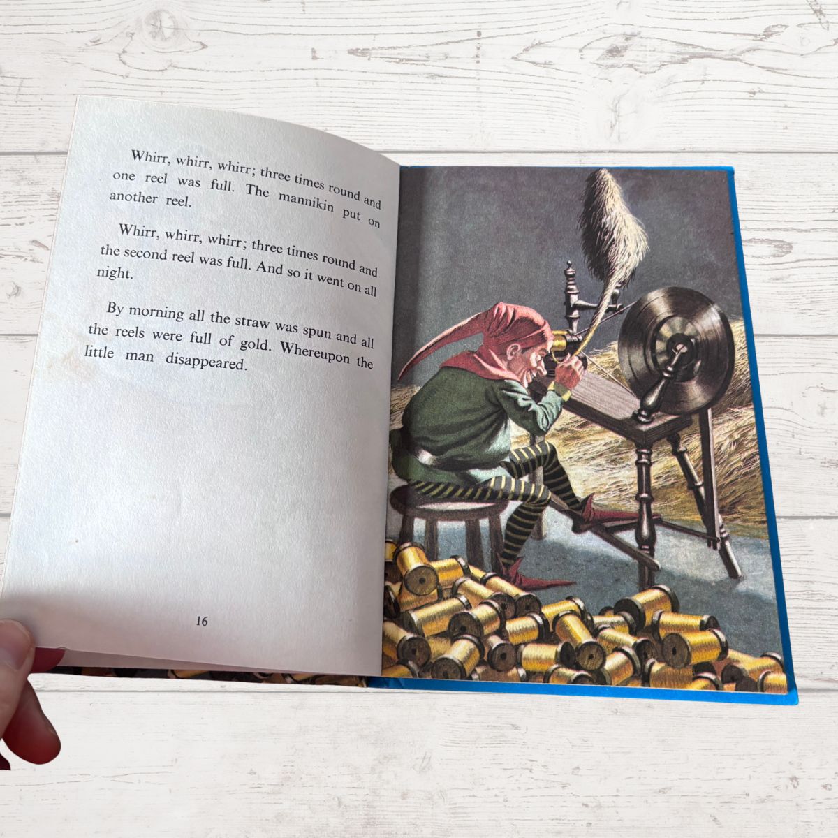 An open children's book lying on a white wood surface, showing a printed story with text on the left page and a colorful illustration on the right page. A hand is visible holding the book open at the lower left corner.