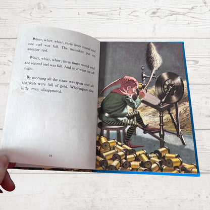 An open children's book lying on a white wood surface, showing a printed story with text on the left page and a colorful illustration on the right page. A hand is visible holding the book open at the lower left corner.