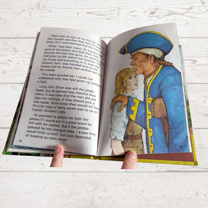 An open vintage hardcover book showing printed text on the left page and a colorful illustration of a pirate on the right page, held open by two hands against a whitewashed wood background.