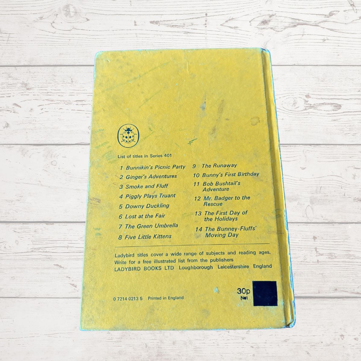 A yellow Ladybird children's activity book, showing the back cover with a list of 14 activities and a small black square sticker, photographed on a white wood background.