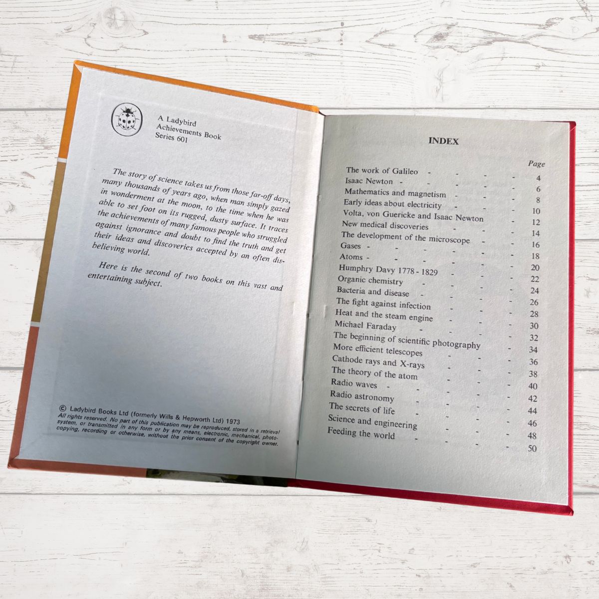 An open vintage book showing printed interior pages with an index, photographed on a light wood surface. The book has a tan cover and red cloth binding visible along the edges.