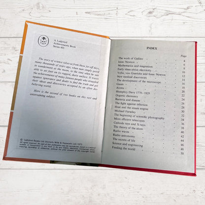 An open vintage book showing printed interior pages with an index, photographed on a light wood surface. The book has a tan cover and red cloth binding visible along the edges.