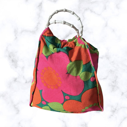 A retro-style bag  with a bold mid-century floral pattern in pink, orange, green, and red, featuring a silver bamboo handle, set against a white marble background.