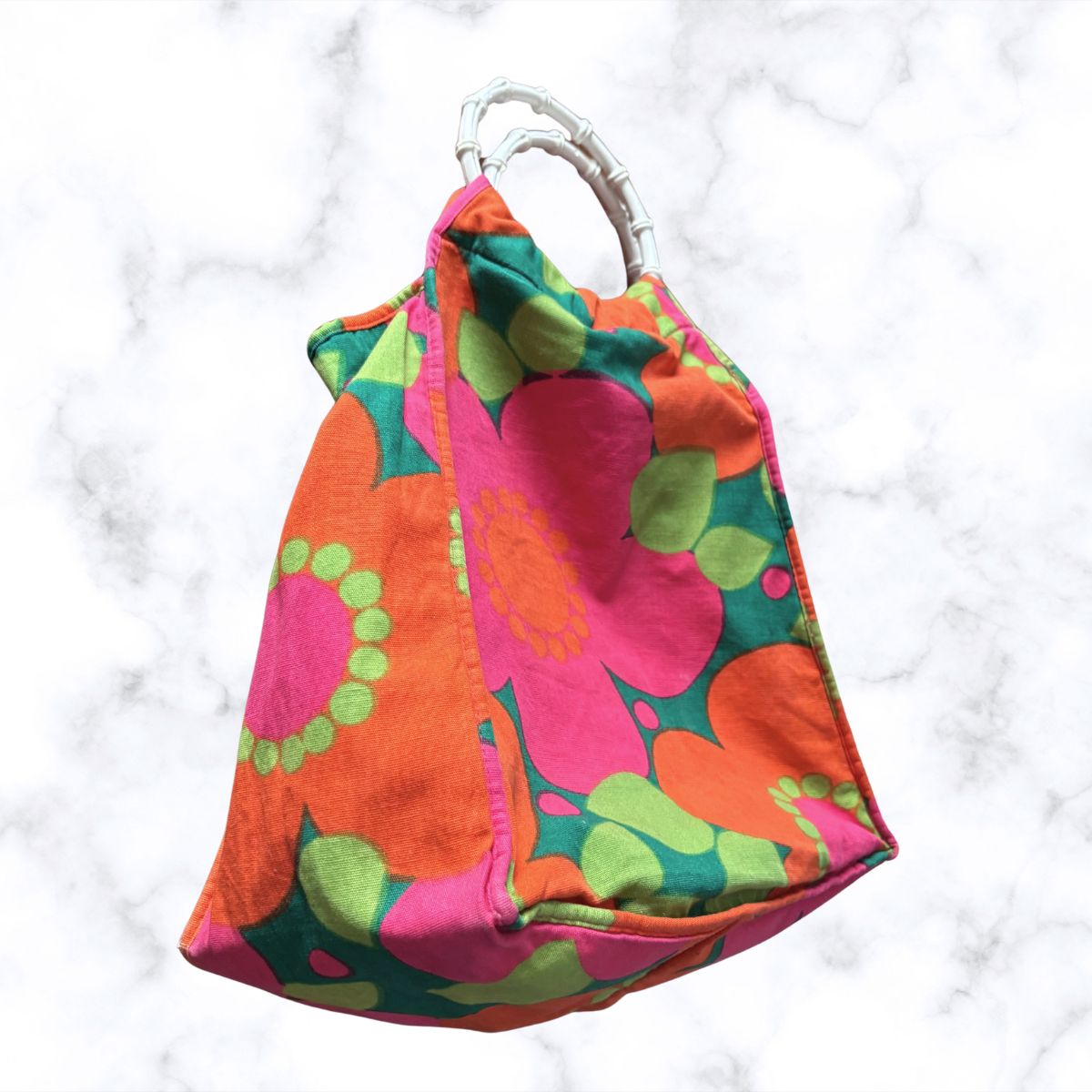 A retro-style fabric tote bag with a bold floral pattern in pink, orange, green, and teal, featuring a silver bamboo-style top handle, set against a white marble background.