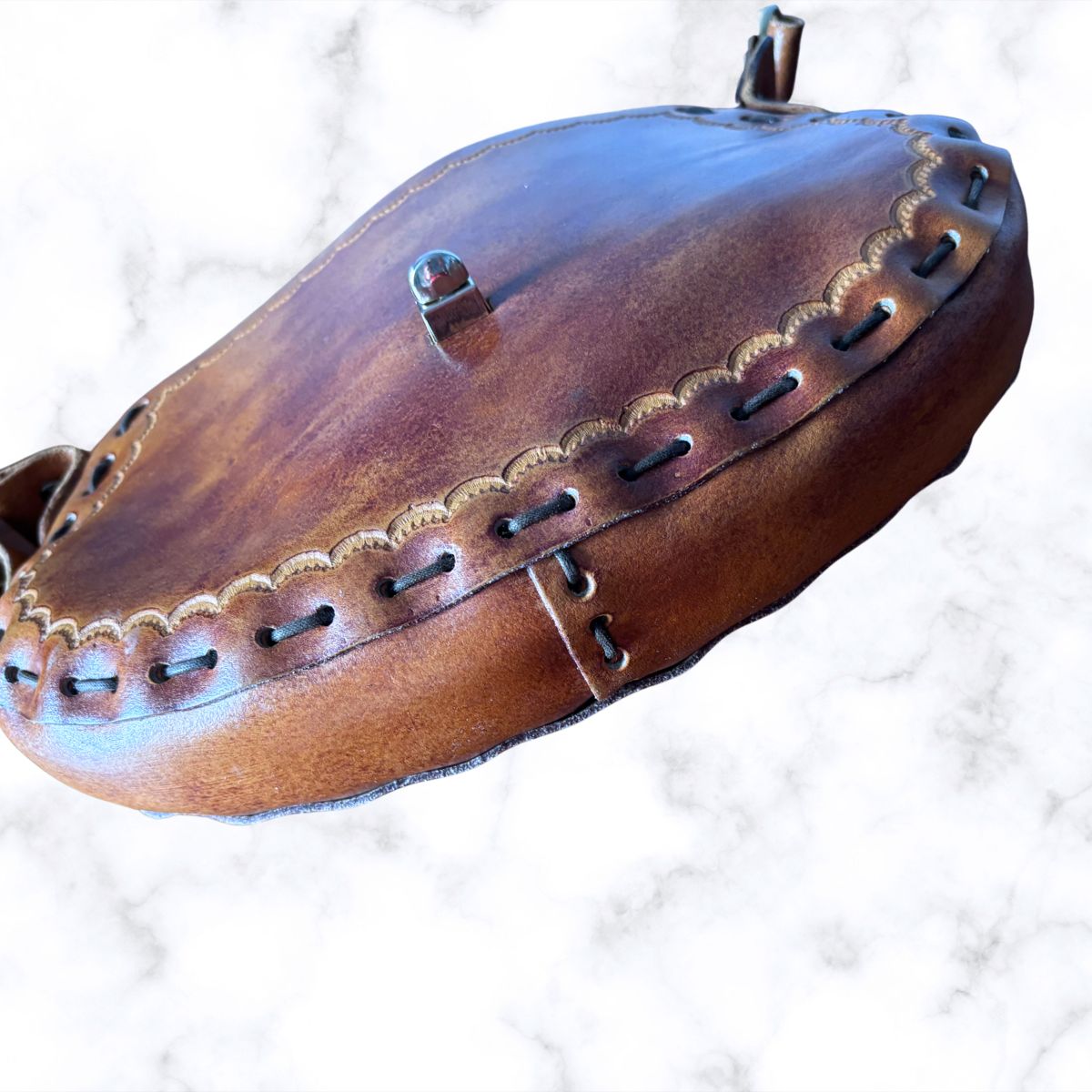 A brown leather bag with decorative stitching and metal hardware, photographed on a white marble-patterned background.