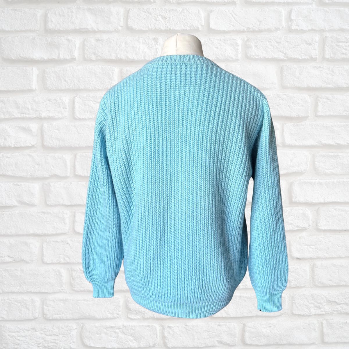 A light blue ribbed knit sweater displayed on a white mannequin torso against a white brick wall background.