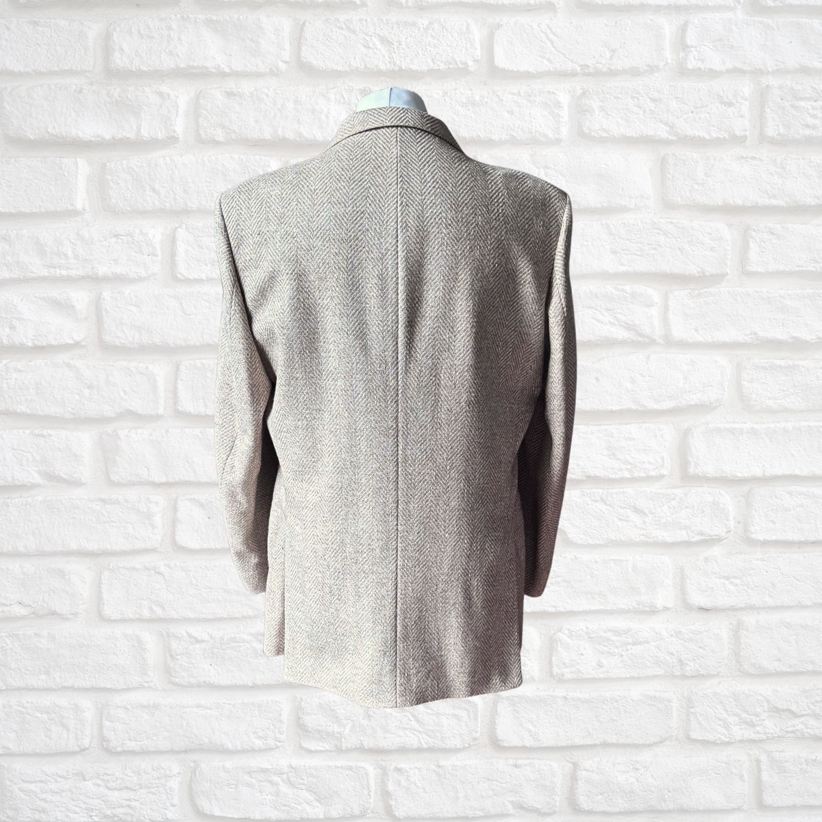 An oatmeal  herringbone tweed blazer shown from the back on a mannequin torso, set against a white brick wall background.