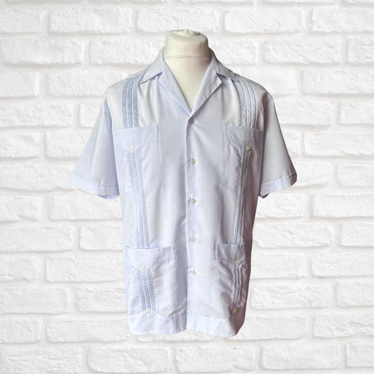 A white short-sleeve guayabera-style shirt displayed on a mannequin torso against a white painted brick wall. The shirt has decorative vertical pleats and button details on the chest and front pockets.