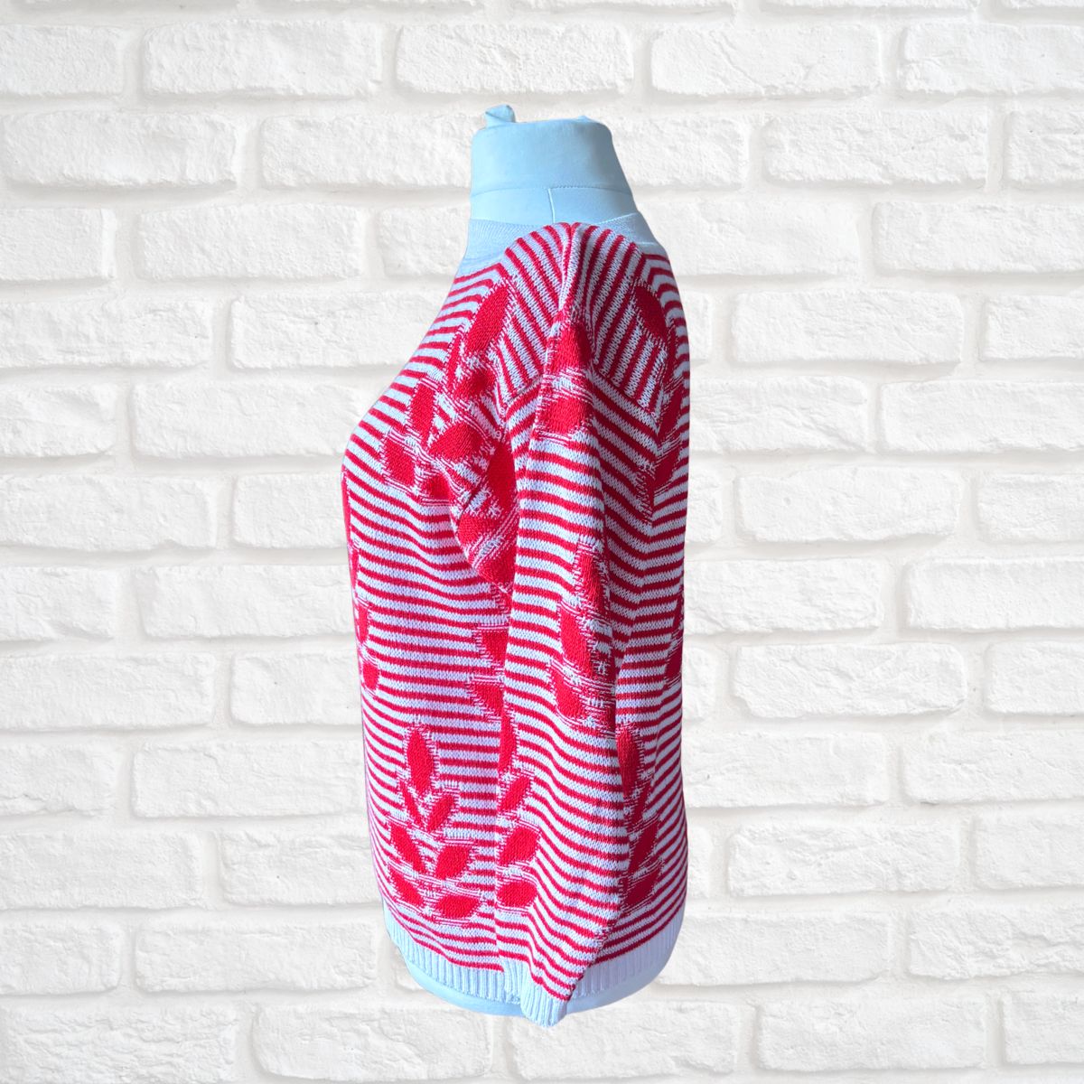 A red and white striped knit sweater with pink floral patterns and light blue ribbed trim, displayed on a light blue mannequin torso against a white brick wall background.