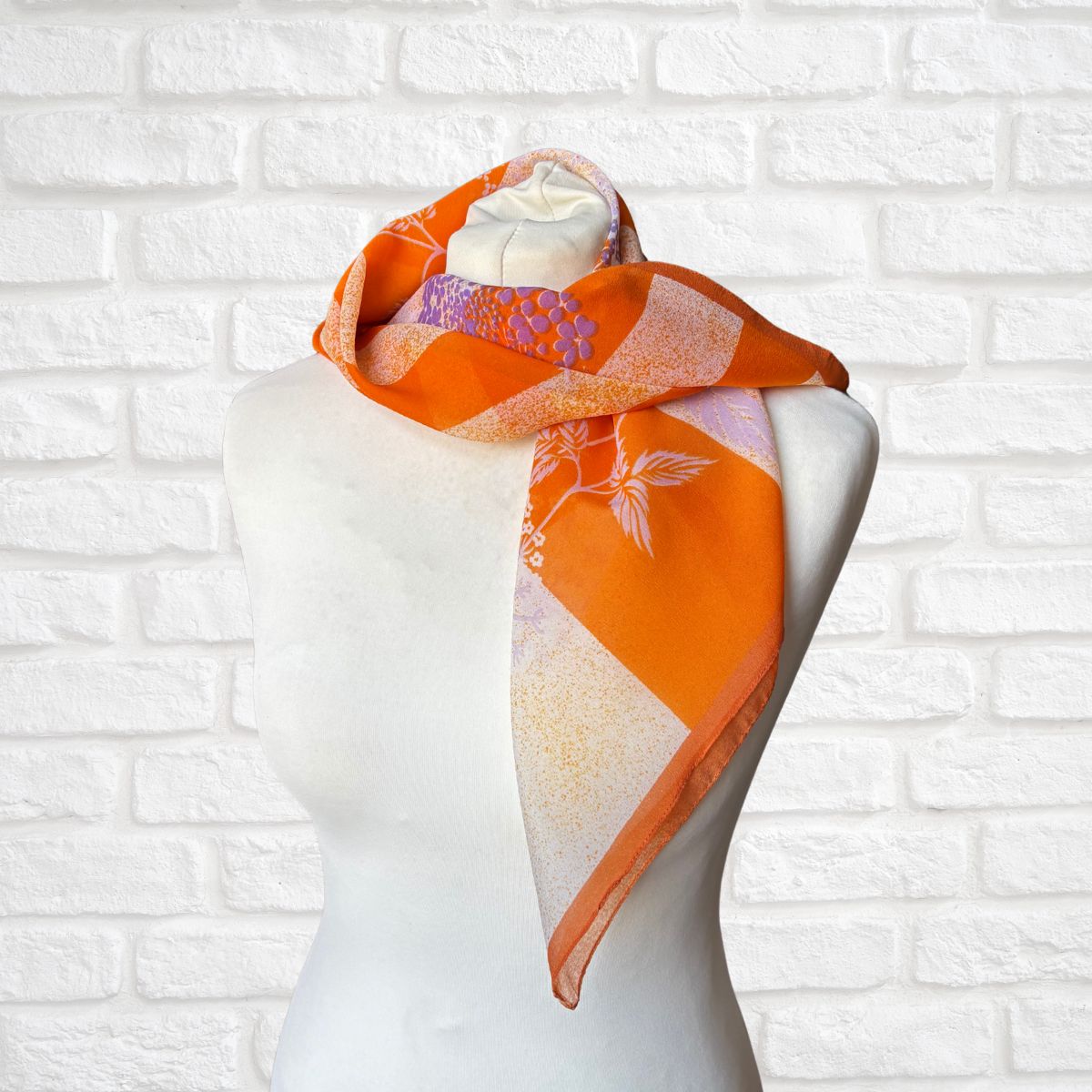 Orange scarf with floral patterns draped over a mannequin against a white brick wall.