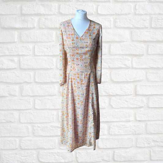 A long-sleeve midi dress with a V-neckline, displayed on a mannequin torso against a white brick wall. The dress features a dense, small-scale multicolor pattern of various cartoon-like characters in orange, yellow, blue, and green on a light beige base.