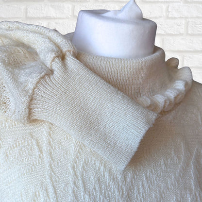 An off-white knit sweater with a ruffled mock neck and textured knit body, displayed on a white mannequin torso against a white brick wall background.