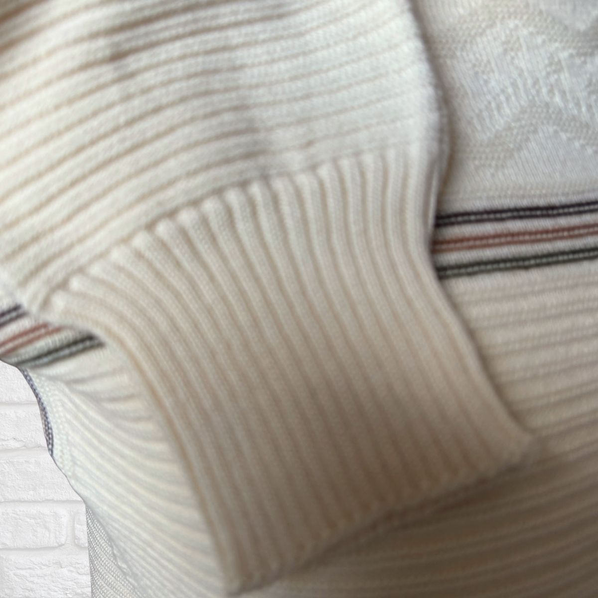 Close-up of a cream-colored ribbed knit sweater sleeve with a striped pattern of thin lines visible in the background.