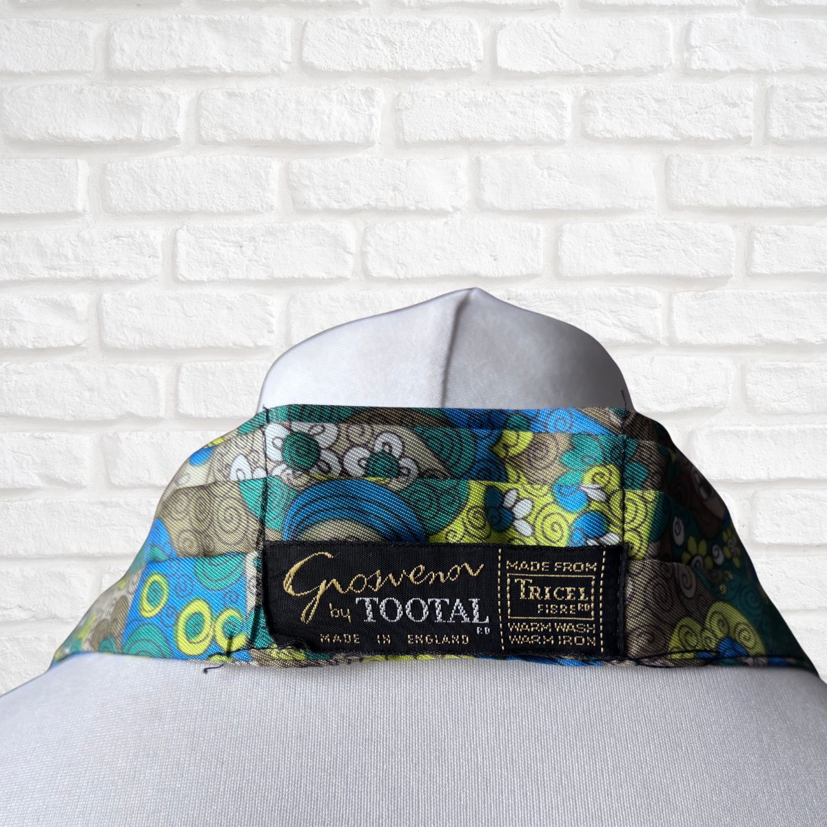 Colorful patterned cravat  on a white shirt with 'Grosvenor by Footal' label against a white brick wall.