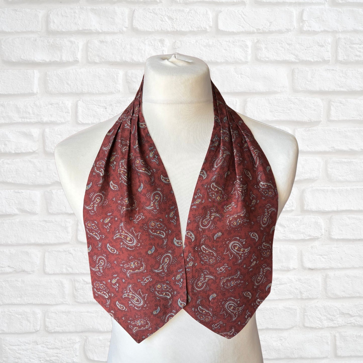 A vintage silk cravat featuring a paisley print in red, grey, yellow, and blue, displayed on a mannequin against a white brick wall background.