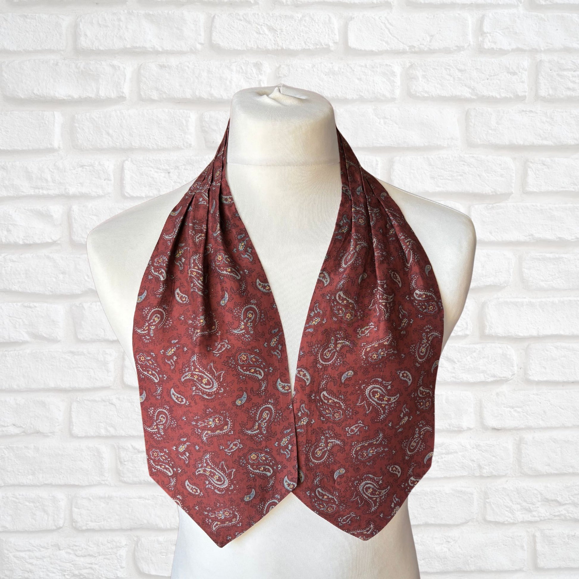 A vintage silk cravat featuring a paisley print in red, grey, yellow, and blue, displayed on a mannequin against a white brick wall background.