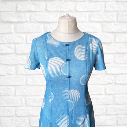 Blue dress with white pattern and toggle bodice detail on a mannequin against a white brick wall.