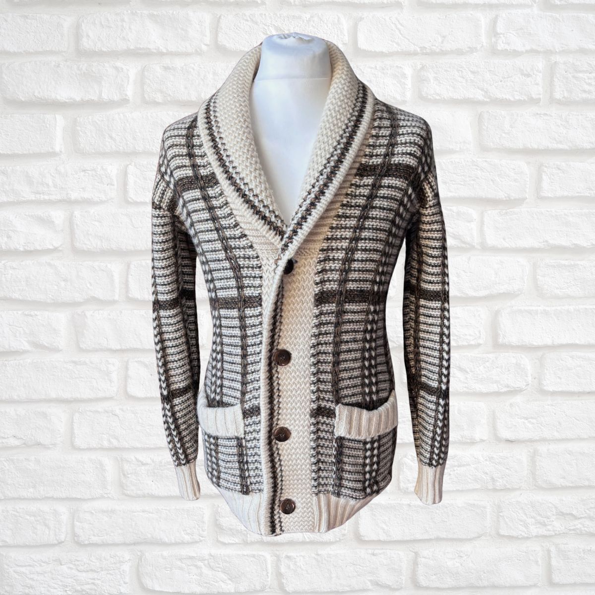 A knit cardigan sweater with a shawl collar, button-front closure, and a plaid pattern, displayed on a white mannequin torso against a white brick wall background.