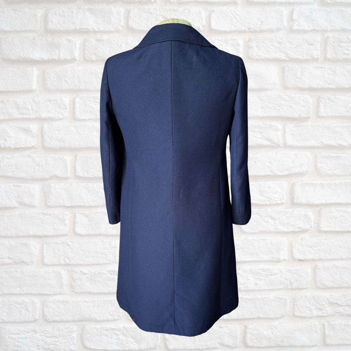 A navy blue longline blazer or coat displayed on a mannequin torso, shown from the back against a white painted brick wall.