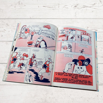 An open vintage comic book laid flat on a white wood surface. The pages show black-and-white cartoon panels with red headline text and pink-toned page backgrounds.