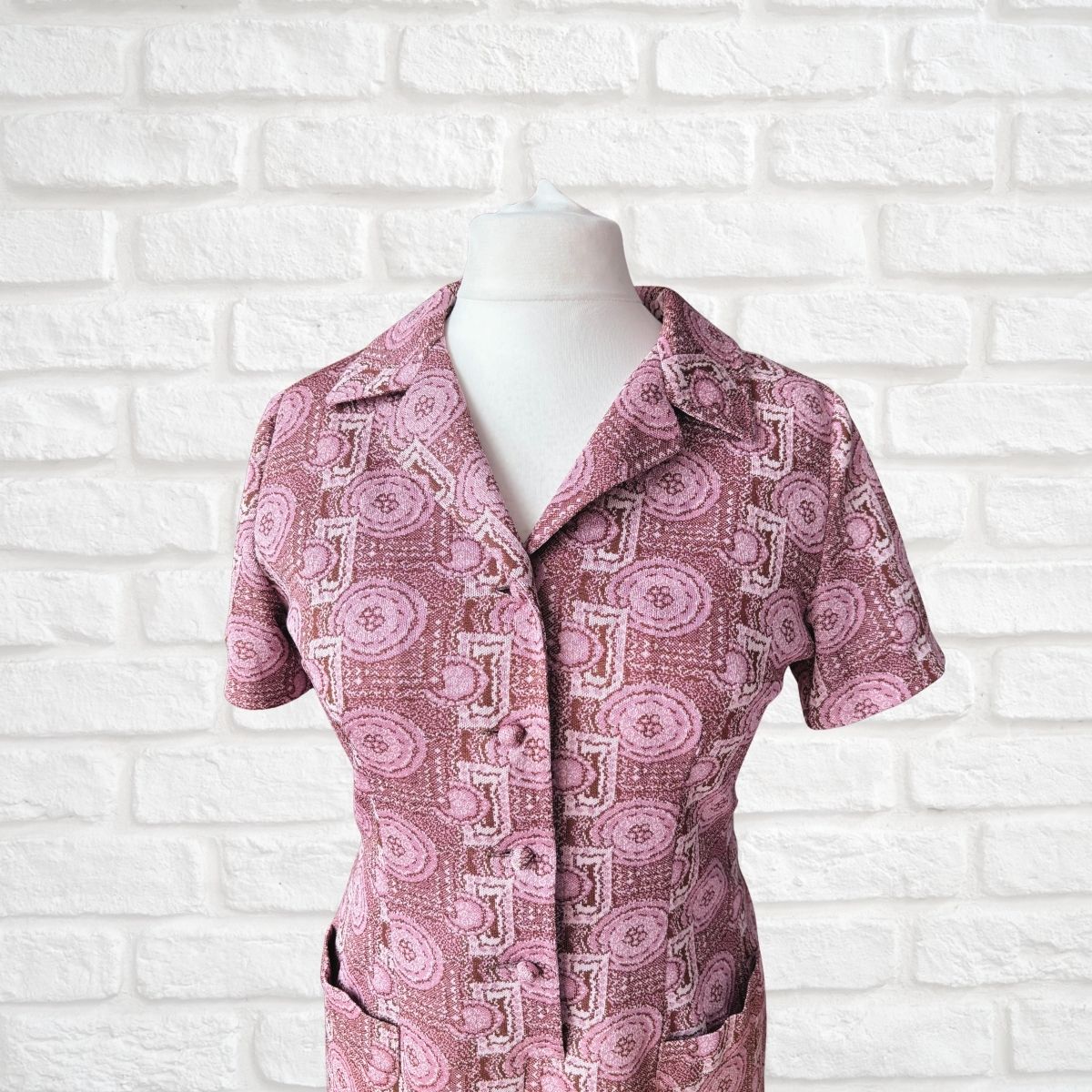 A short-sleeve button-front shirt dress in dusty pink and white with a circular medallion pattern, displayed on a white mannequin torso against a white brick wall background.