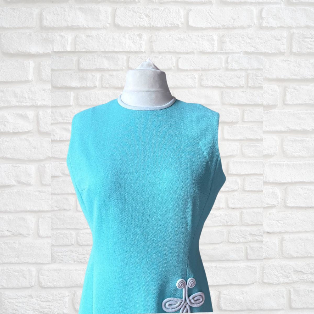 Turquoise sleeveless  dress with white embroidery on a mannequin against a white brick wall.