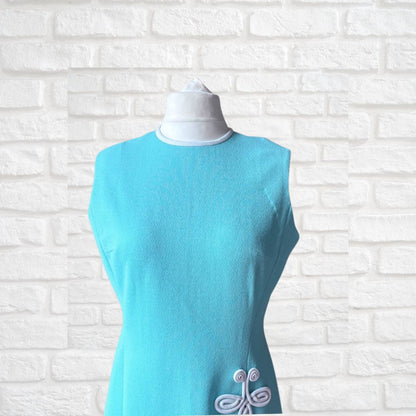 Turquoise sleeveless  dress with white embroidery on a mannequin against a white brick wall.
