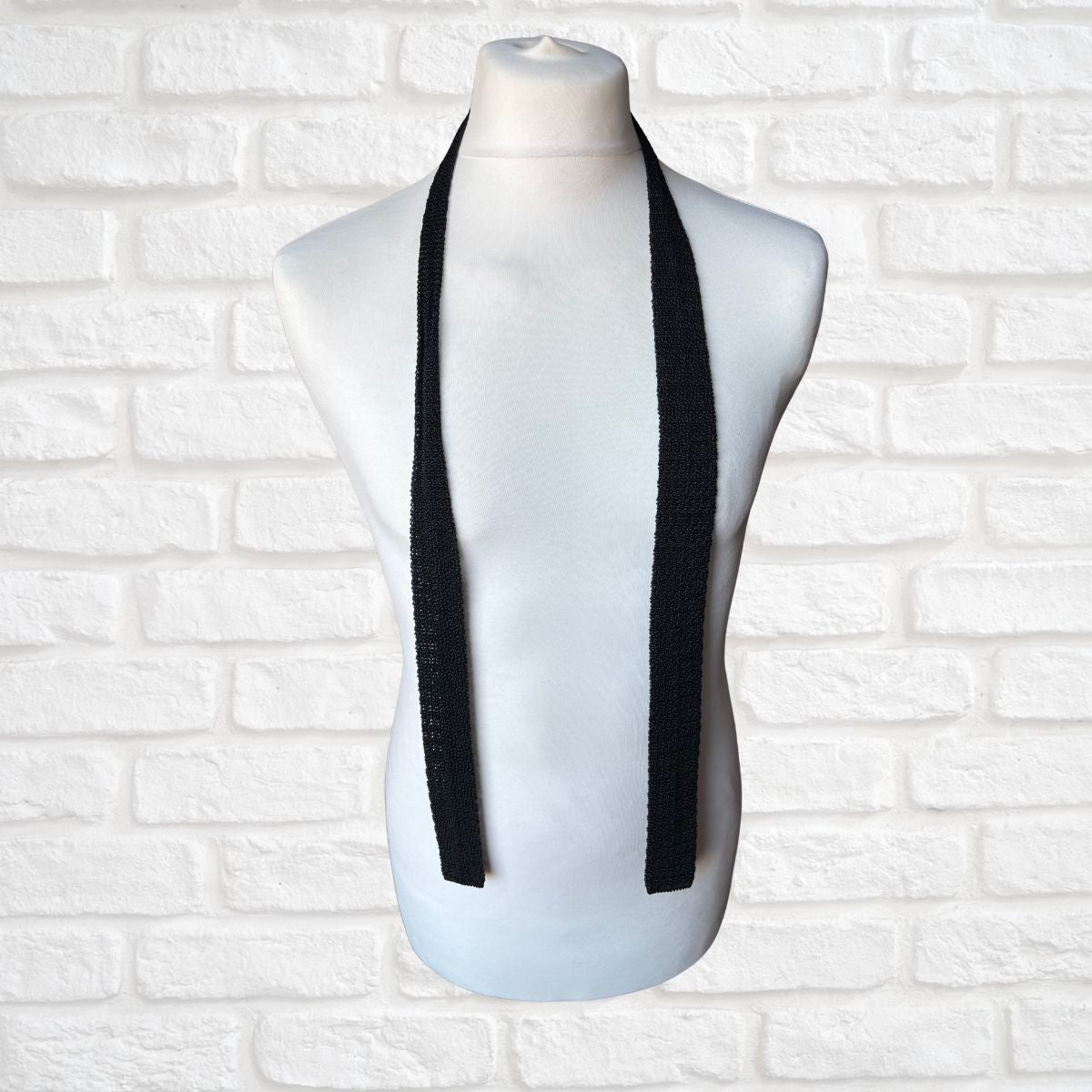 Black 60s skinny tie on a mannequin bust against a white brick wall background