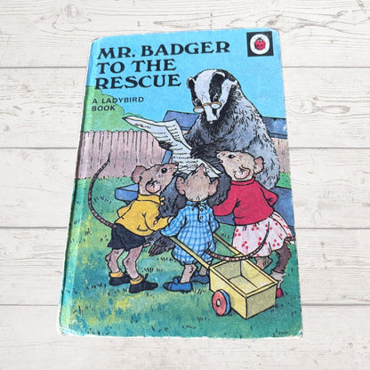 Vintage hardcover book titled 'Mr. Badger to the Rescue' by Ladybird, featuring an illustration of a badger and three mice with a small yellow cart, placed on a white wooden surface.