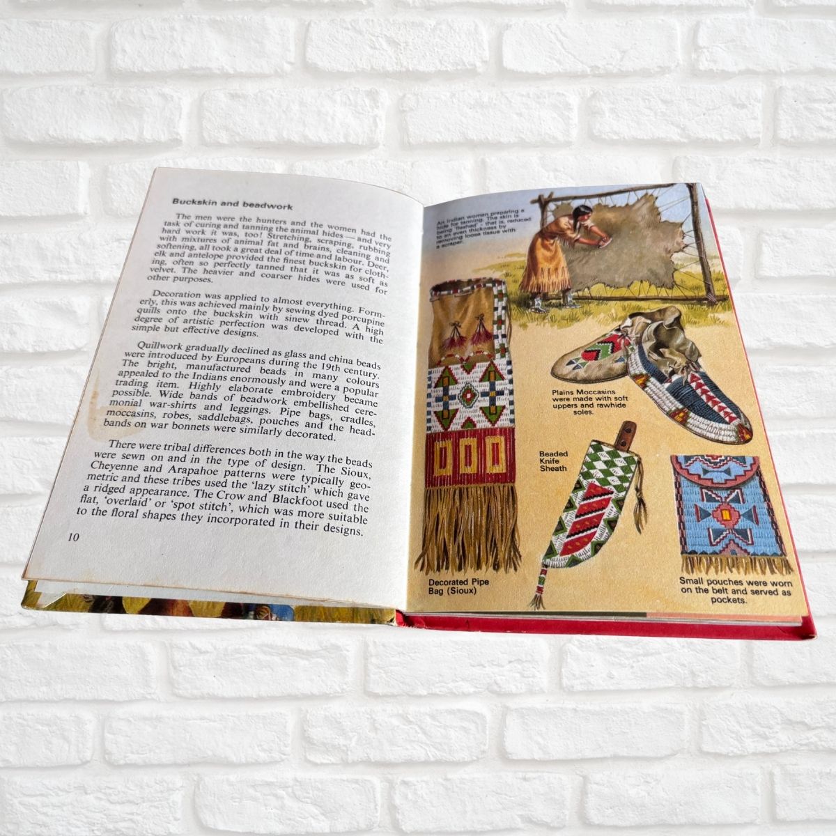 An open book showing pages with text and illustrations of Native American beaded garments and a moccasin, photographed against a white brick wall background.