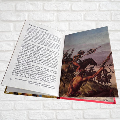 An open book spread displayed on a white brick wall, showing printed text on the left page and a historical battle illustration on the right page with Native American figures on horseback.